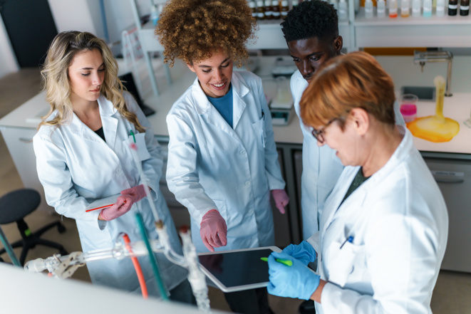 Gruppe von unterschiedlichen jungen Studenten der Naturwissenschaften und ihrem Professor, die während eines Experiments in einem modernen Labor zusammenarbeiten, ein digitales Tablet benutzen und Notizen machen